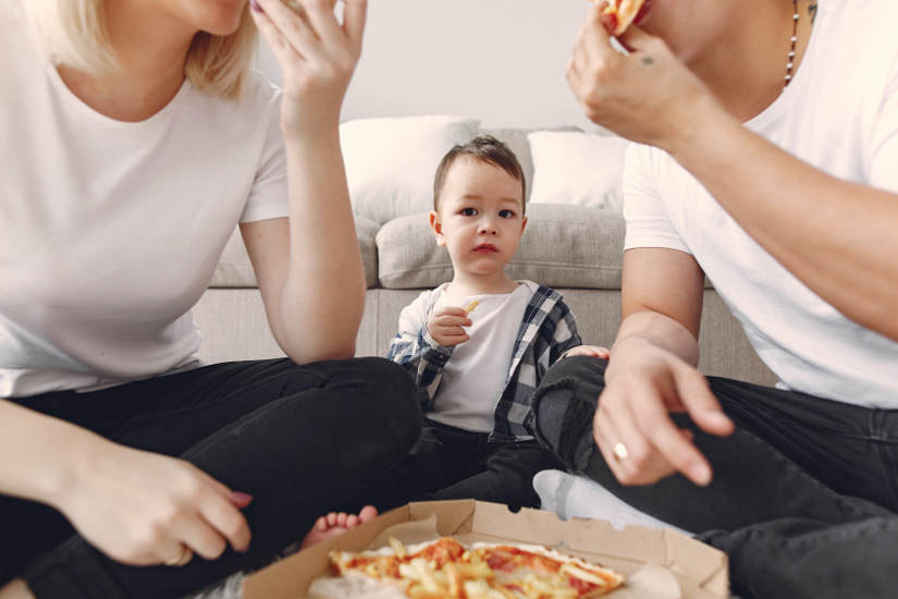 Kid holding finger food looks on as parents enjoy pizza